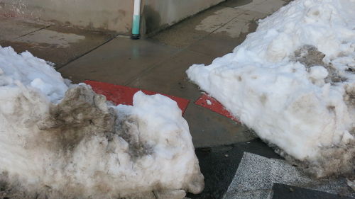 Snow piled up at a street corner, with a narrow path dug out through it.