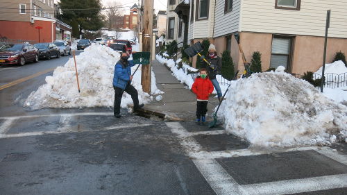 A street corner with a path shoveled through the snow pile. Three people who helped shovel out the crosswalk pose for a photo.