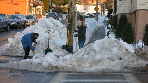 Snow piled up at a street corner. Two people are shoveling the snow out of the crosswalk.