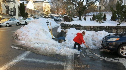 Snow piled up at a street corner. Two people are shoveling snow from the crosswalk.