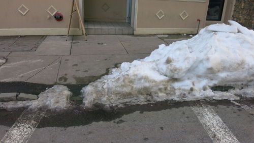 Small mound of packed, frozen snow obstructing the crosswalk curb ramp at an intersection.